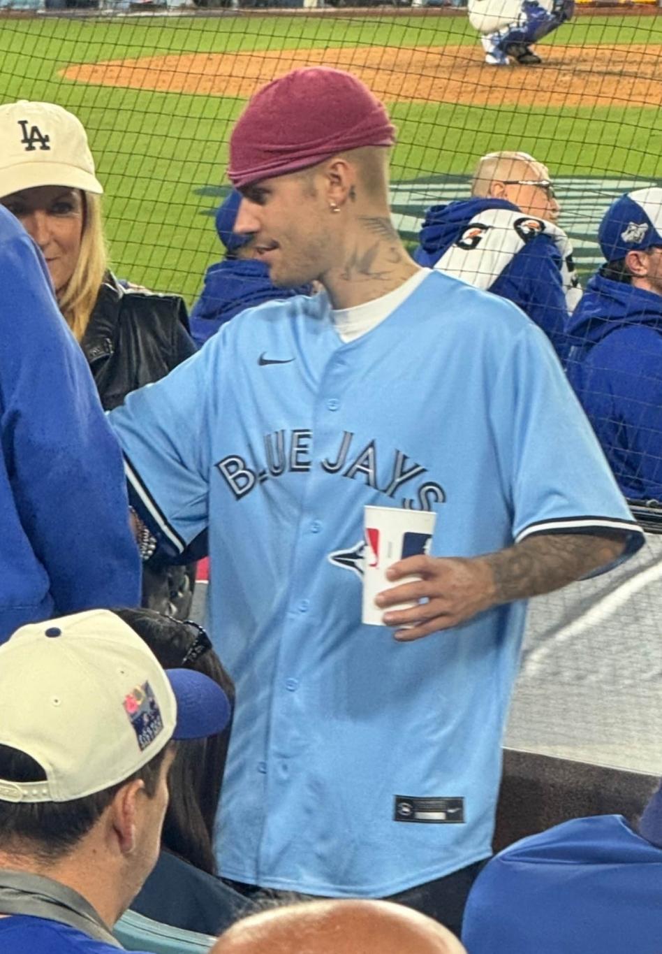 Джастин Бибер в голубой майке "Торонто Блю Джейс" и красной кепке, с напитком на бейсбольном матче.