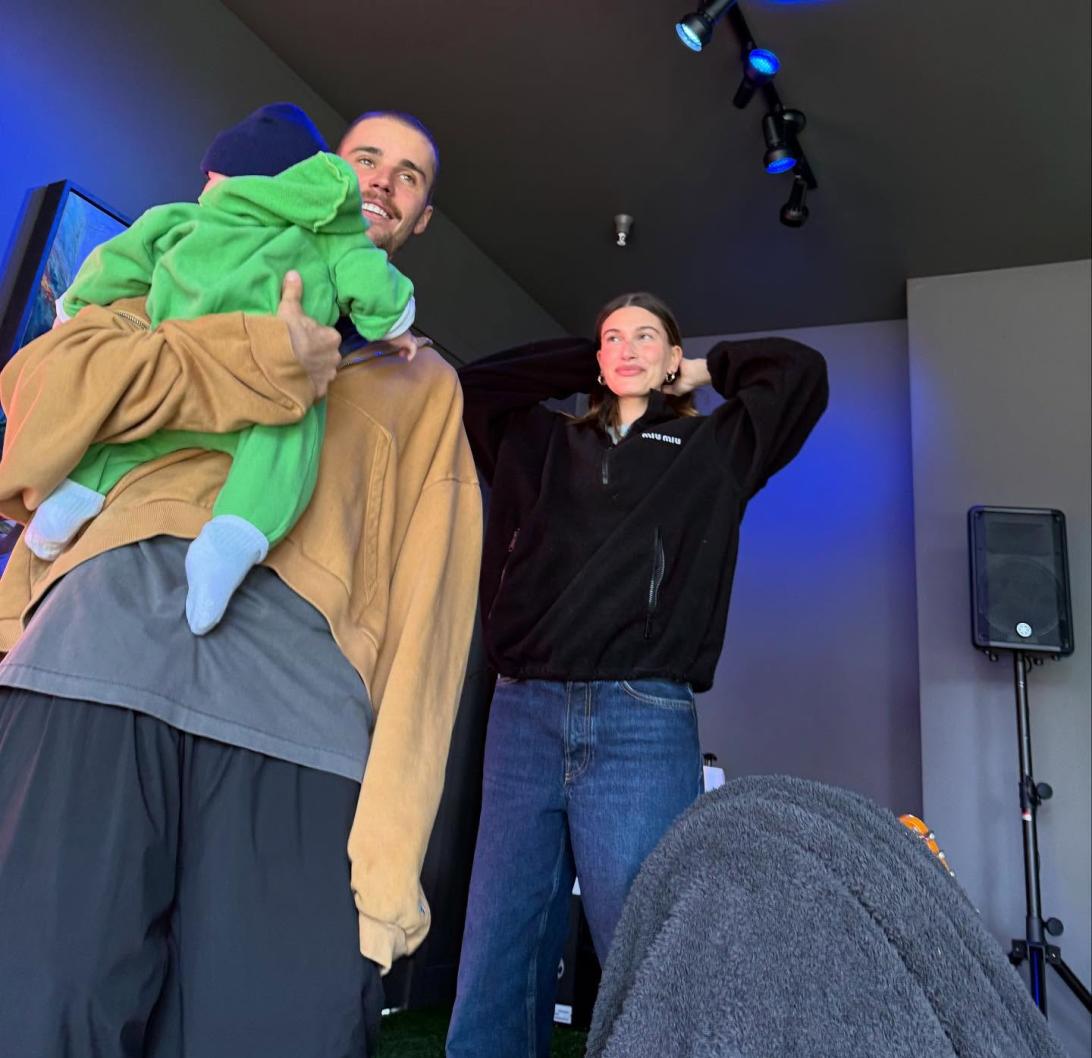 Джастин Бибер держит на руках малыша в зеленом наряде, рядом с ним стоит Хейли Бибер.