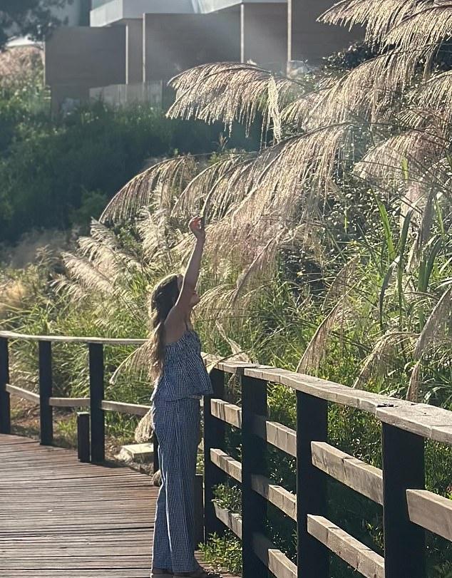 На фото дочь в полосатом летнем комплекте тянется рукой к листьям дерева над головой.