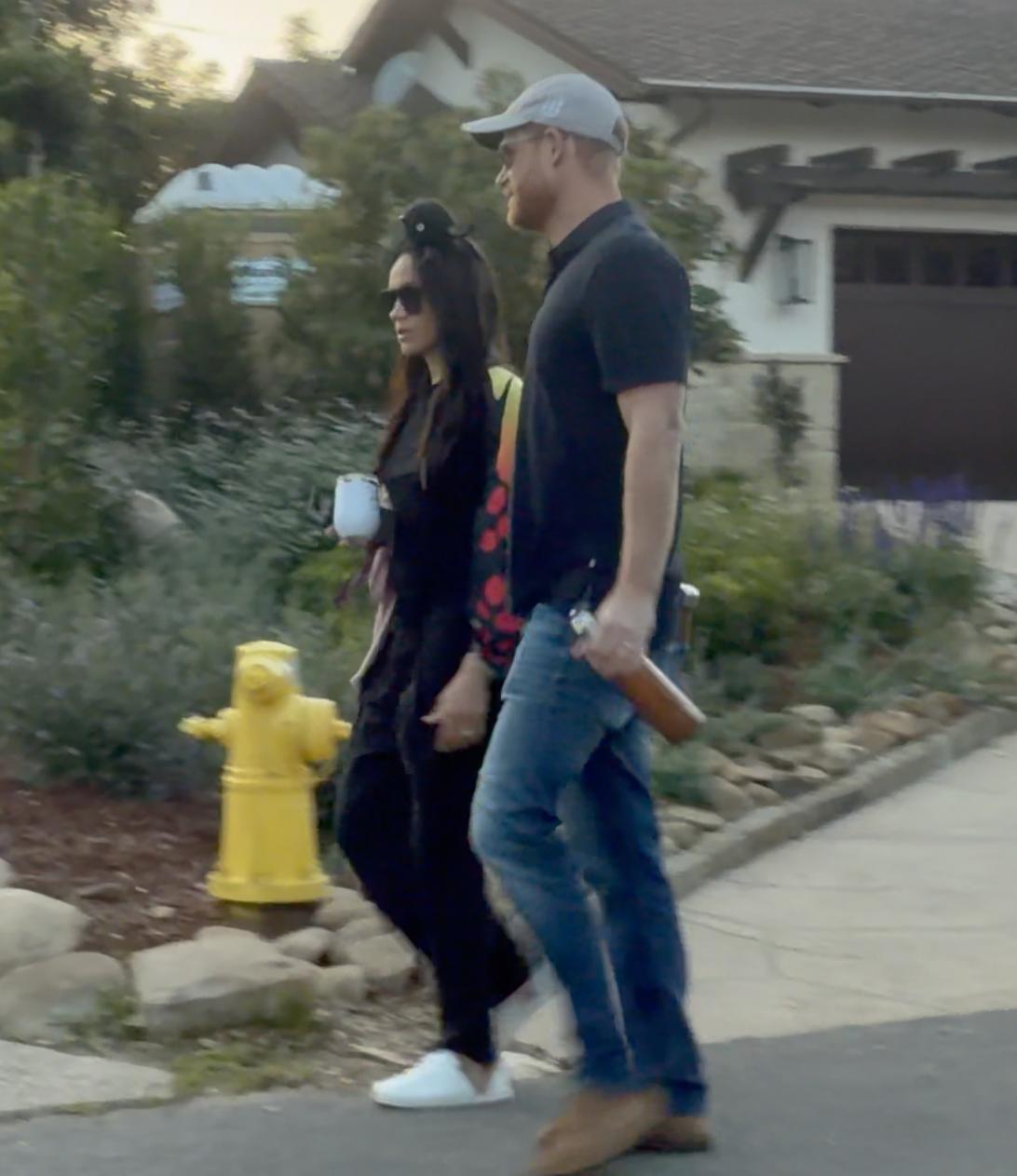 Принц Гарри в кепке с двумя пивными бутылками и Меган Маркл в очках с бокалом вина идут по улице.