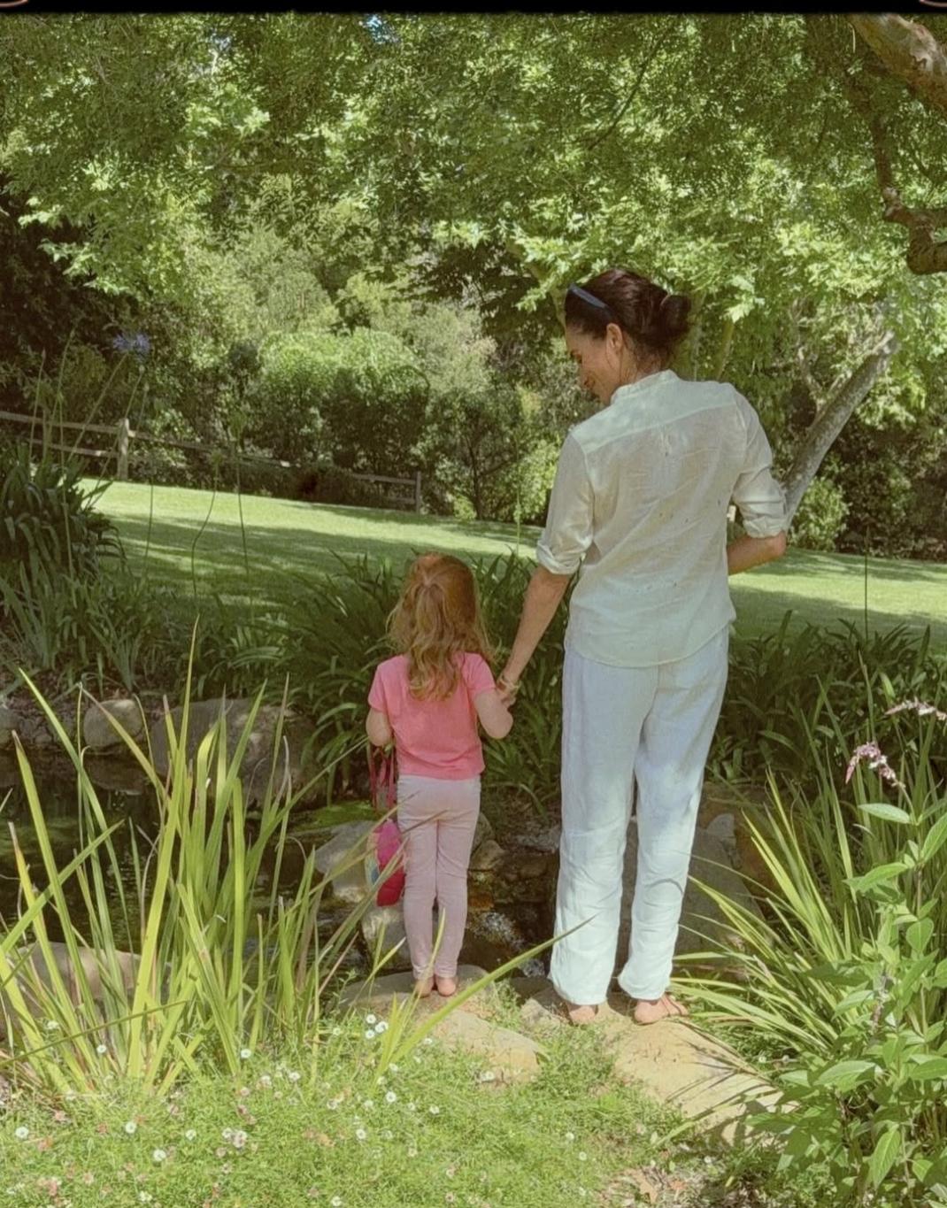 Меган Маркл и принцесса Лилибет стоят босиком, держась за руки, на каменной тропинке у небольшого ручья в пышном саду.