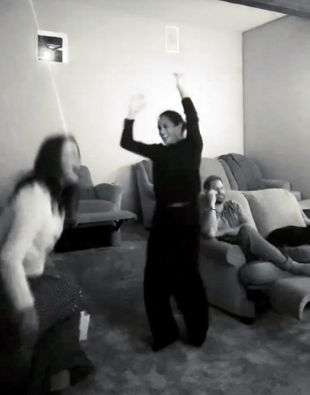 Меган Маркл танцует с другой женщиной на фоне сидящего на диване принца Гарри.