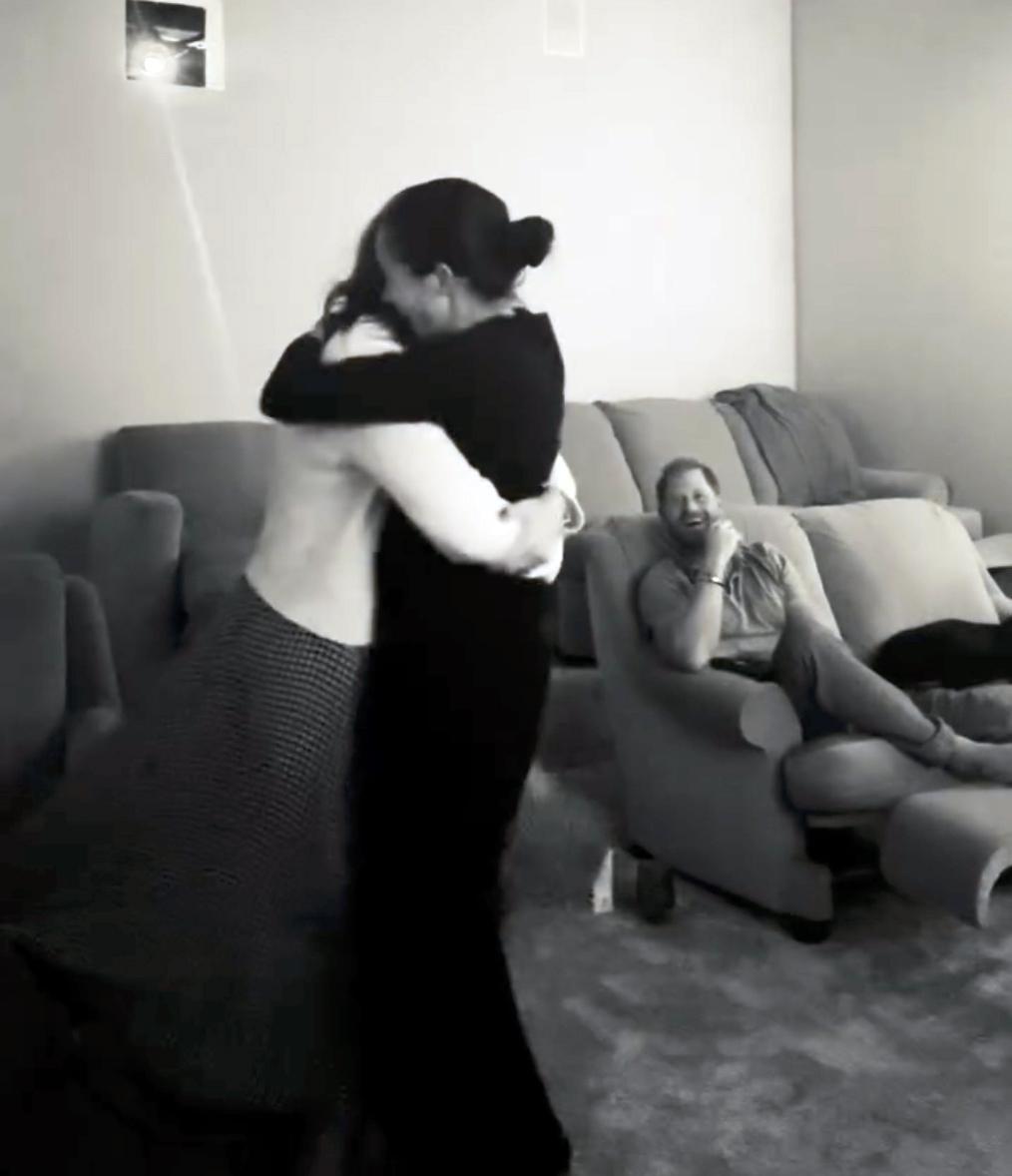 Меган Маркл тепло обнимает женщину, а принц Гарри улыбается на заднем плане.