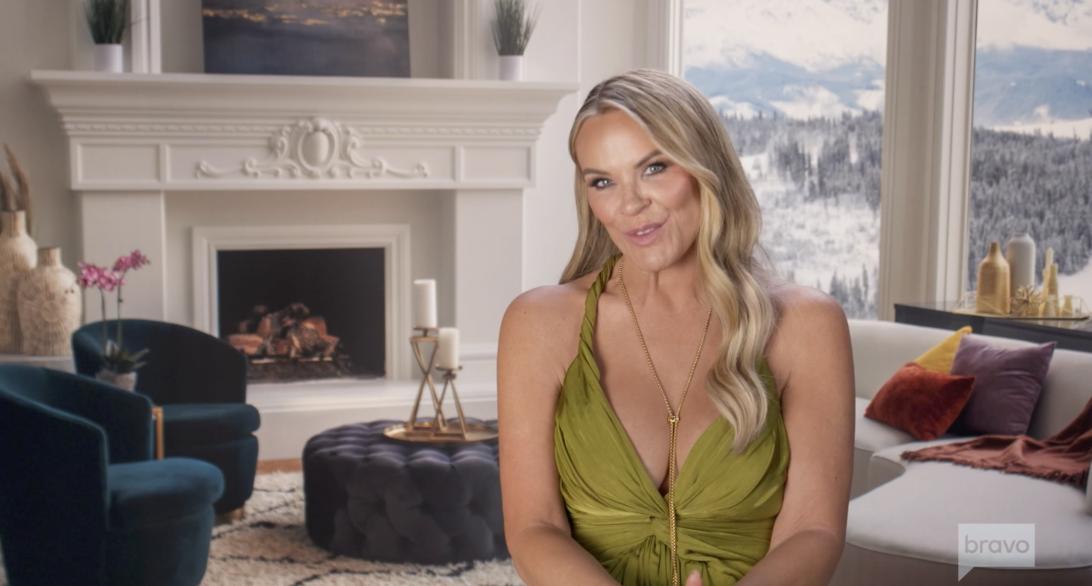 Хизер Гей в зеленом платье и золотом колье позирует в роскошной гостиной с панорамным видом на снежные горы.