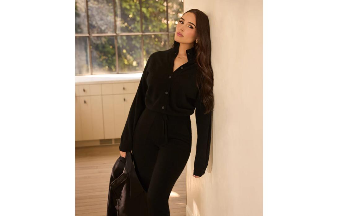 Женщина в черном кардигане и брюках в тон, с черной курткой в руках, прислонилась к стене.