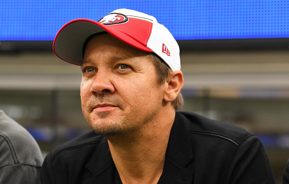 Актер Джереми Реннер в красно-белой кепке смотрит матч НФЛ.