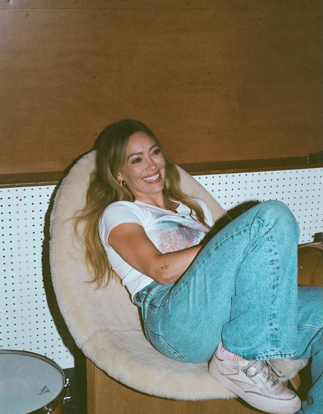 Хилари Дафф улыбается в пушистом кресле, с барабаном на переднем плане, вероятно, в студии звукозаписи.