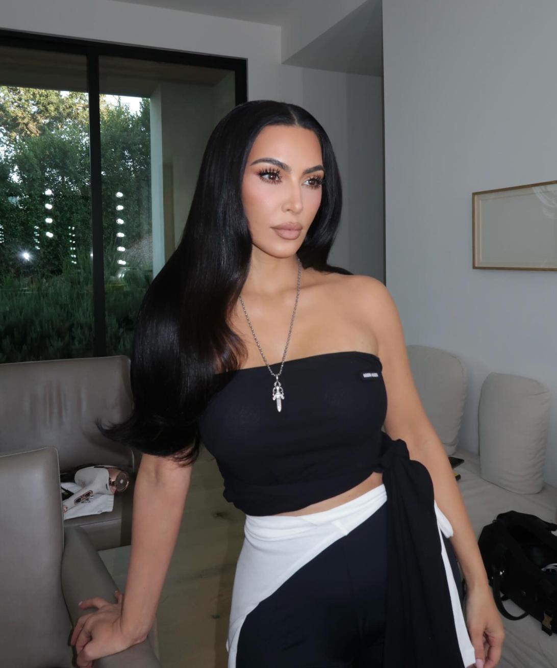 Ким Кардашьян позирует в помещении в черном топе-бандо и двухцветных брюках.