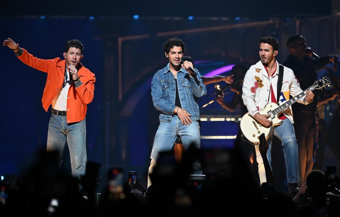 Братья Джонас выступают на сцене: Ник поет, Джо поет, держа микрофон, Кевин играет на белой электрогитаре.