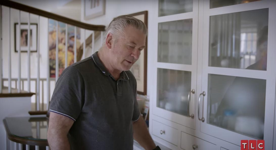 Алек Болдуин стоит на кухне с белыми шкафами, в сером поло.