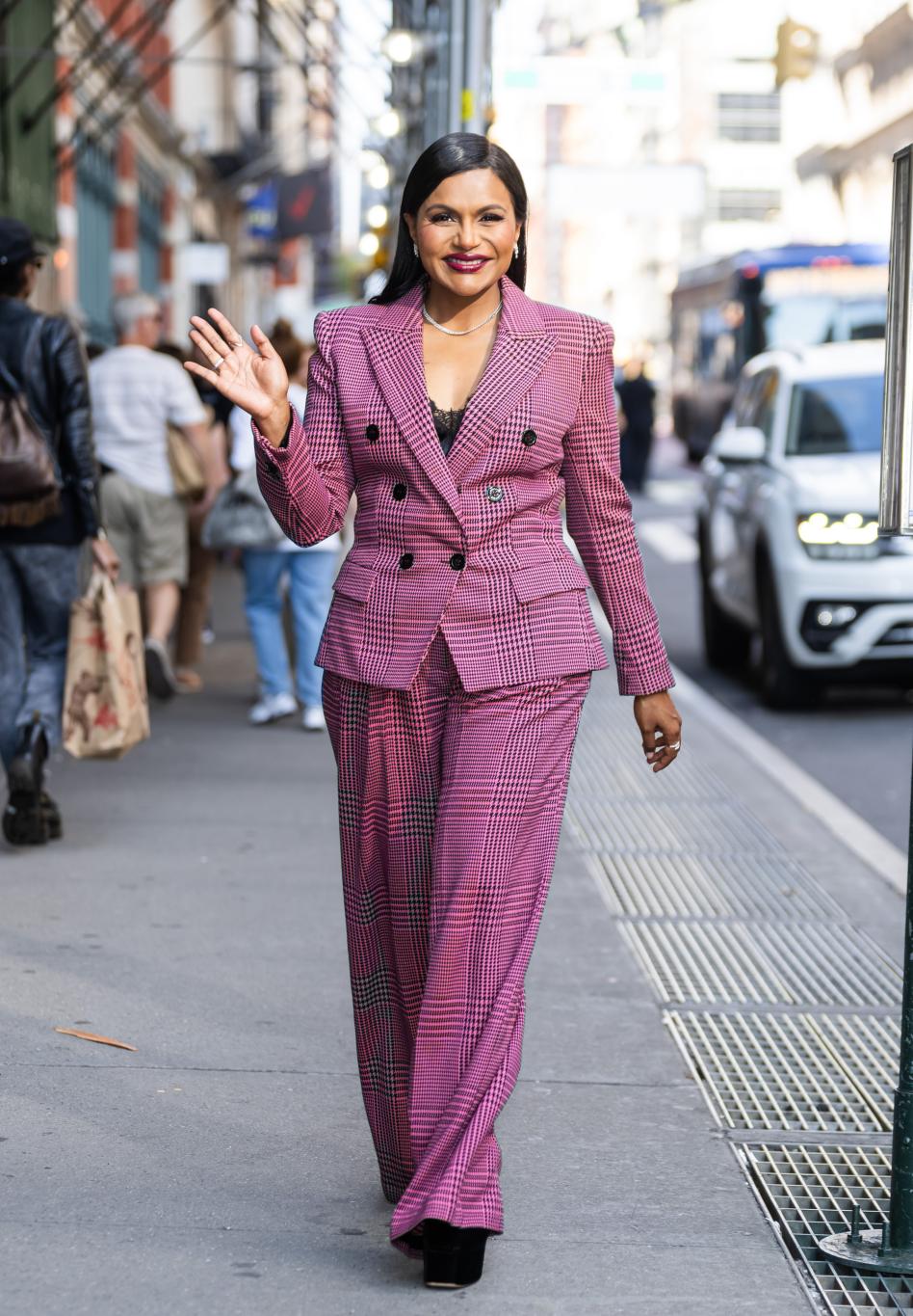 Минди Калинг, улыбающаяся и машущая в розовом клетчатом брючном костюме на городской улице.