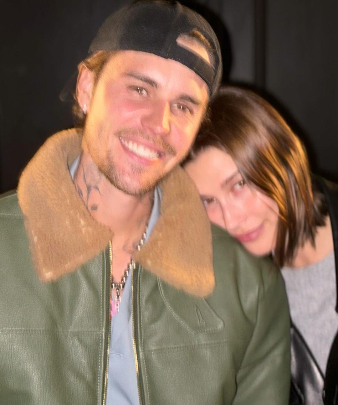 Джастин Бибер улыбается в черной кепке и зеленой кожаной куртке, пока Хейли Бибер нежно прижимается головой к его плечу.