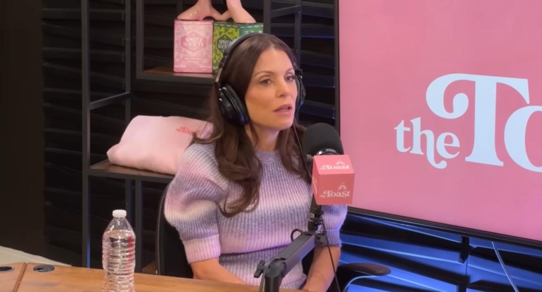 Бетенни Фрэнкел в наушниках и фиолетово-белом полосатом свитере вещает в микрофон с логотипом "The Toast".