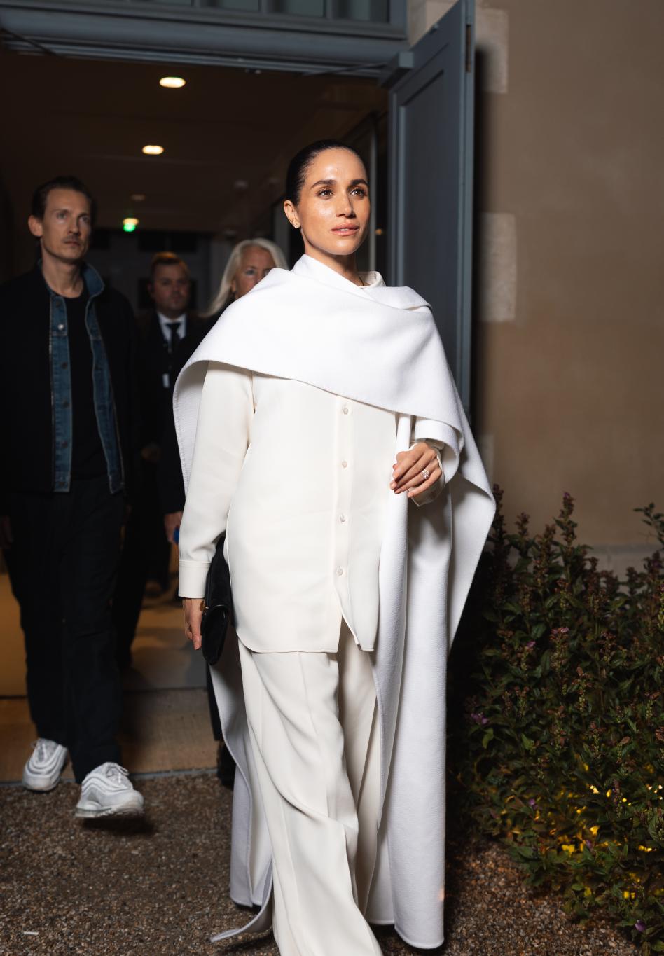 Меган Маркл в белоснежном ансамбле: струящаяся рубашка, широкие брюки, эффектная накидка. Прогулка на улице с гладким пучком и минимумом золота.