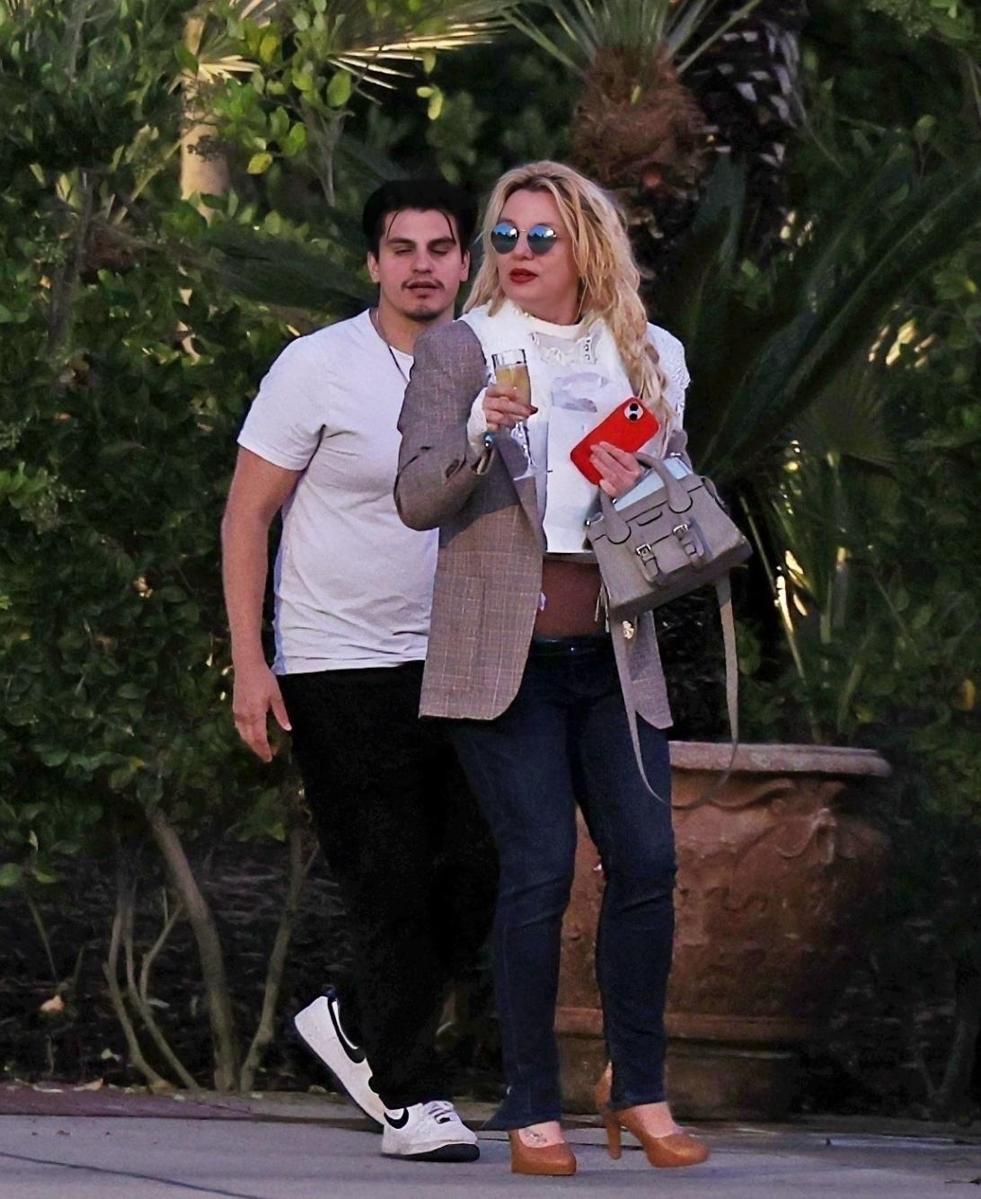 Бритни Спирс в зеркальных очках, с бокалом шампанского, красным телефоном и серой сумочкой, прогуливается с мужчиной.