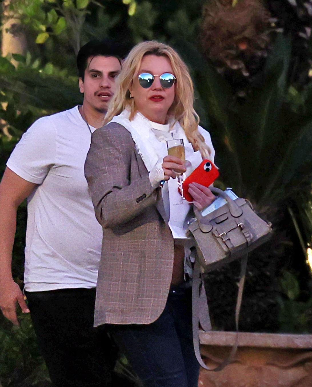 Бритни Спирс держит бокал шампанского и телефон, сумочка на руке, за ней стоит мужчина.