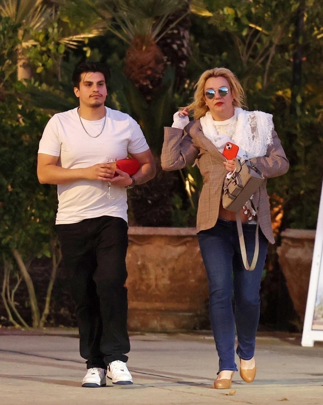 Бритни Спирс с телефоном шагает рядом с сотрудником, несущим бокал шампанского и красную Bluetooth-колонку.