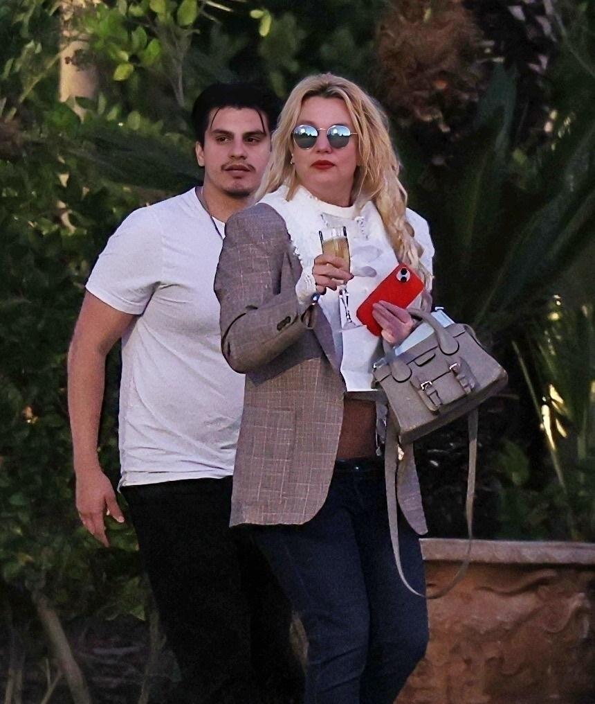 Бритни Спирс с бокалом шампанского и красным телефоном прогуливается на фоне мужчины.