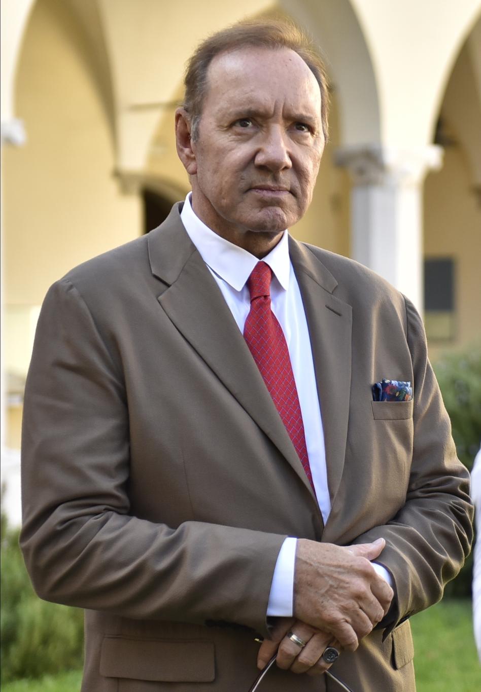 Знаменитый американский актёр Кевин Спейси в коричневом костюме и красном галстуке.