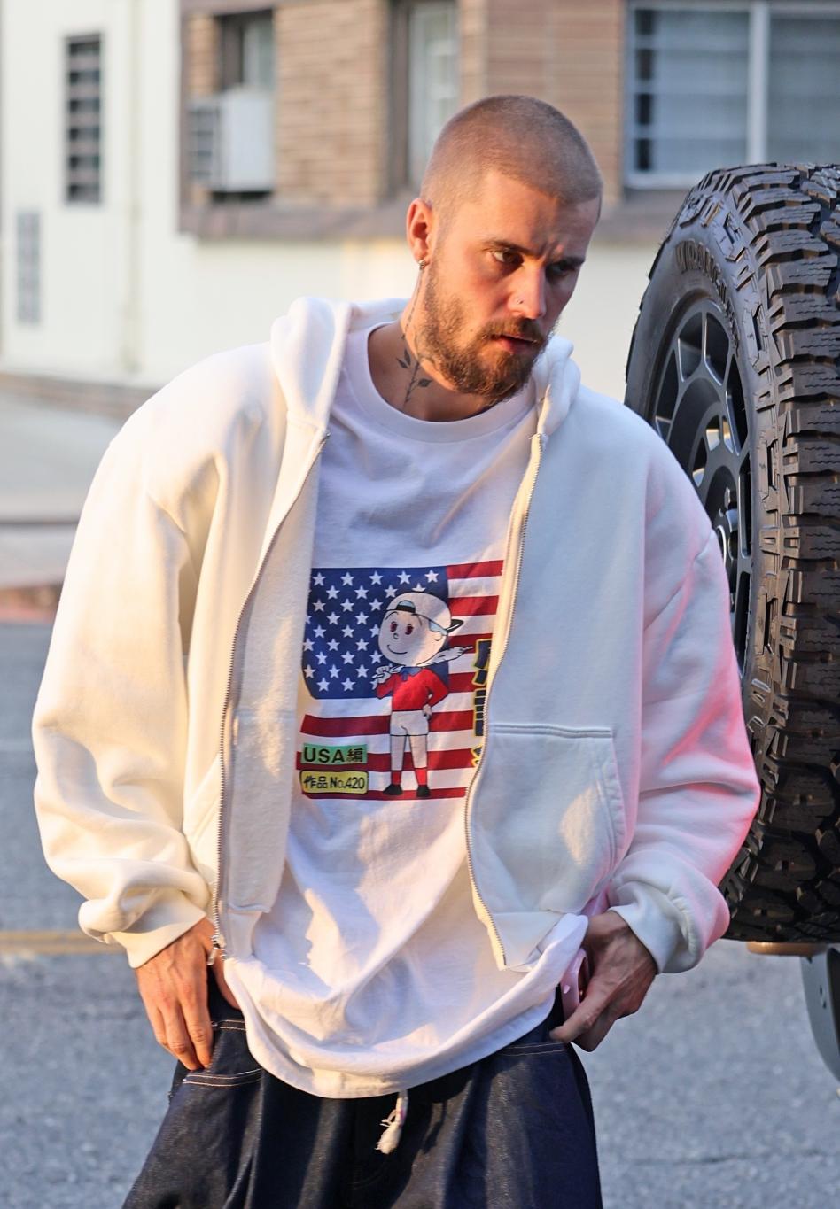 Джастин Бибер в белом худи поверх белой футболки с американским флагом и мультяшным принтом, с грустью опустив взгляд.