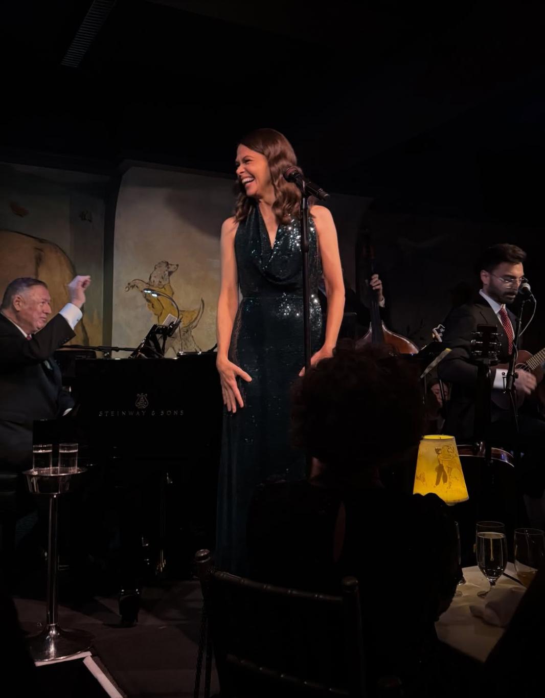 Саттон Фостер выступает на сцене в темно-зеленом платье, расшитом пайетками, а за ней — музыканты с роялем.