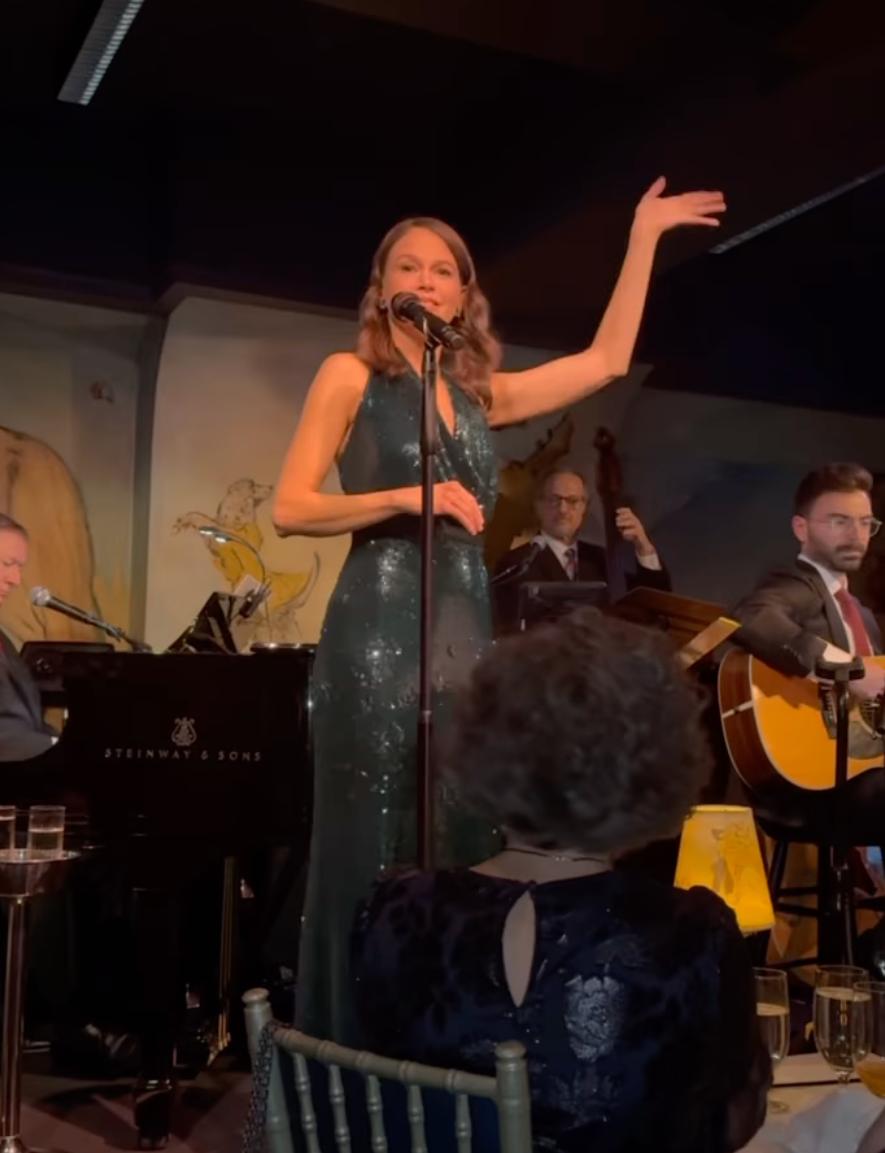 Саттон Фостер, в сияющем зеленом платье с пайетками, исполняет песню у микрофона, в окружении музыкантов.