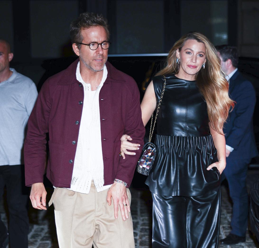 Райан Рейнольдс в бордовой куртке и Блейк Лайвли в черном кожаном платье с узорчатой сумочкой шагают под руку.