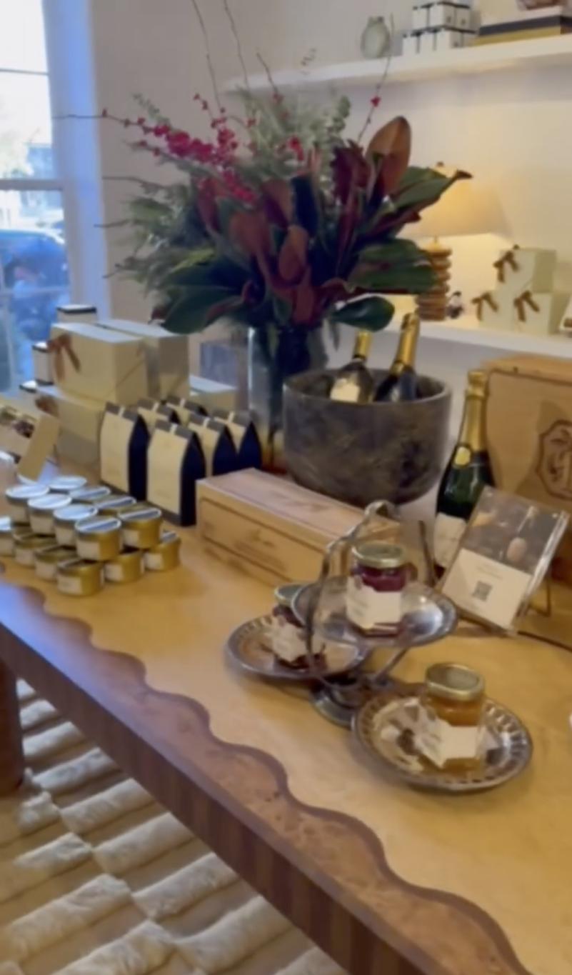 Деревянный стол, богато уставленный: завернутые подарки, небольшие баночки, бутылки шампанского и цветочная композиция.