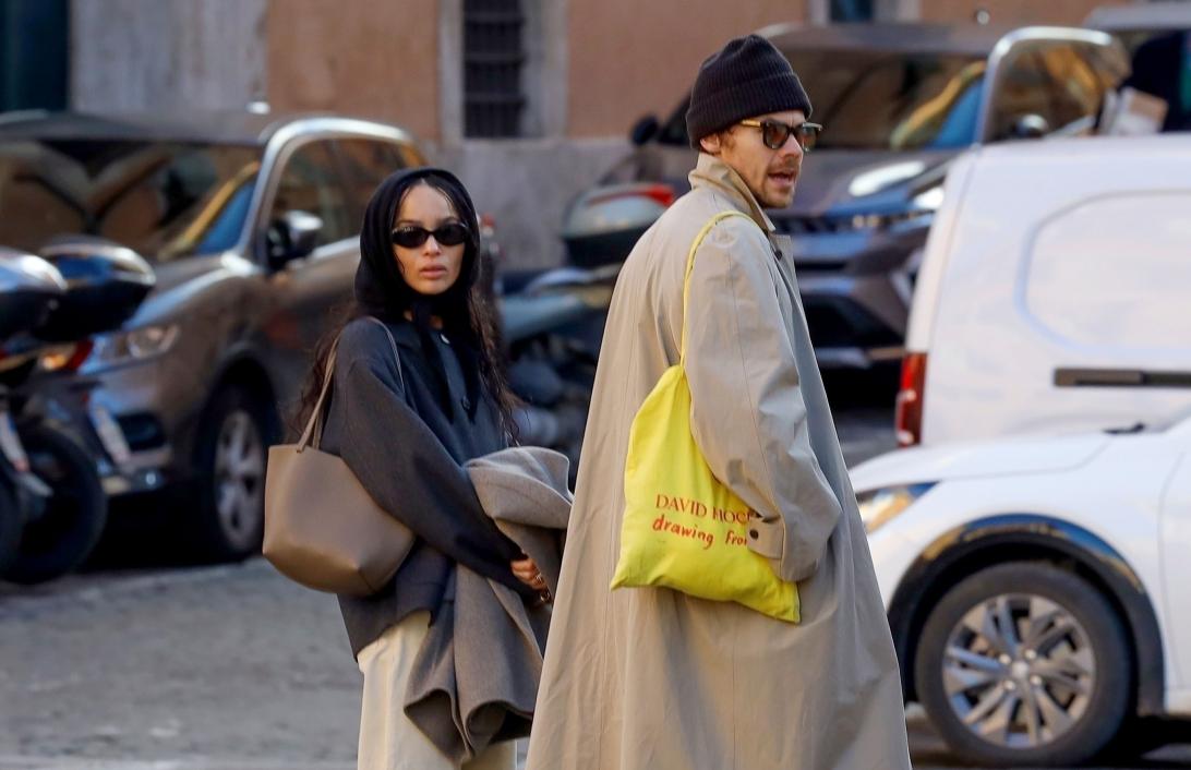 Идут Гарри Стайлс и Зои Кравиц: он в бежевом тренче с желтой сумкой-шопером, она в черном худи и бежевых брюках.