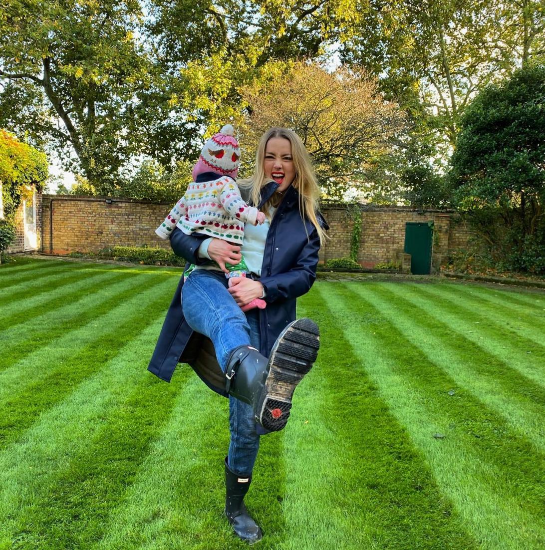 Эмбер Херд с малышом на руках, задорно подняв ногу на лужайке.