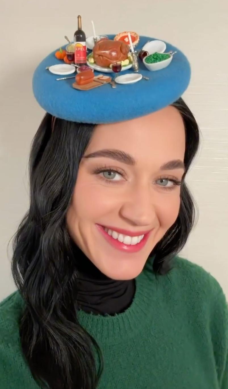 Улыбающаяся Кэти Перри в синей шляпе, украшенной миниатюрными блюдами Дня Благодарения: индейкой, вином и пирогом.