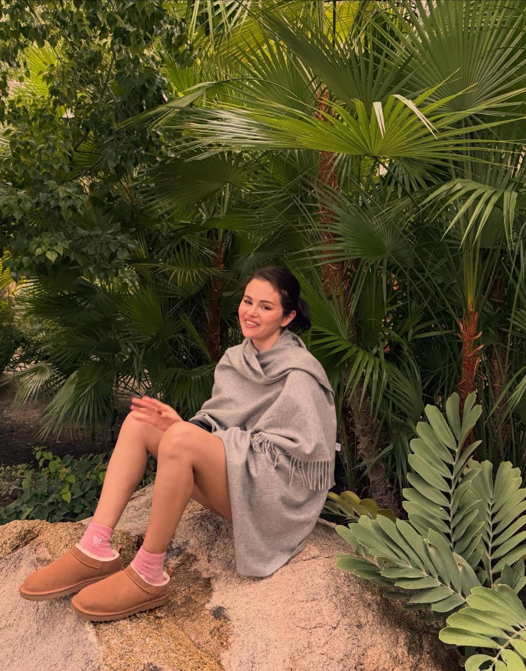 Улыбающаяся Селена Гомес сидит на камне, укутанная серым пледом, среди буйной зелени.