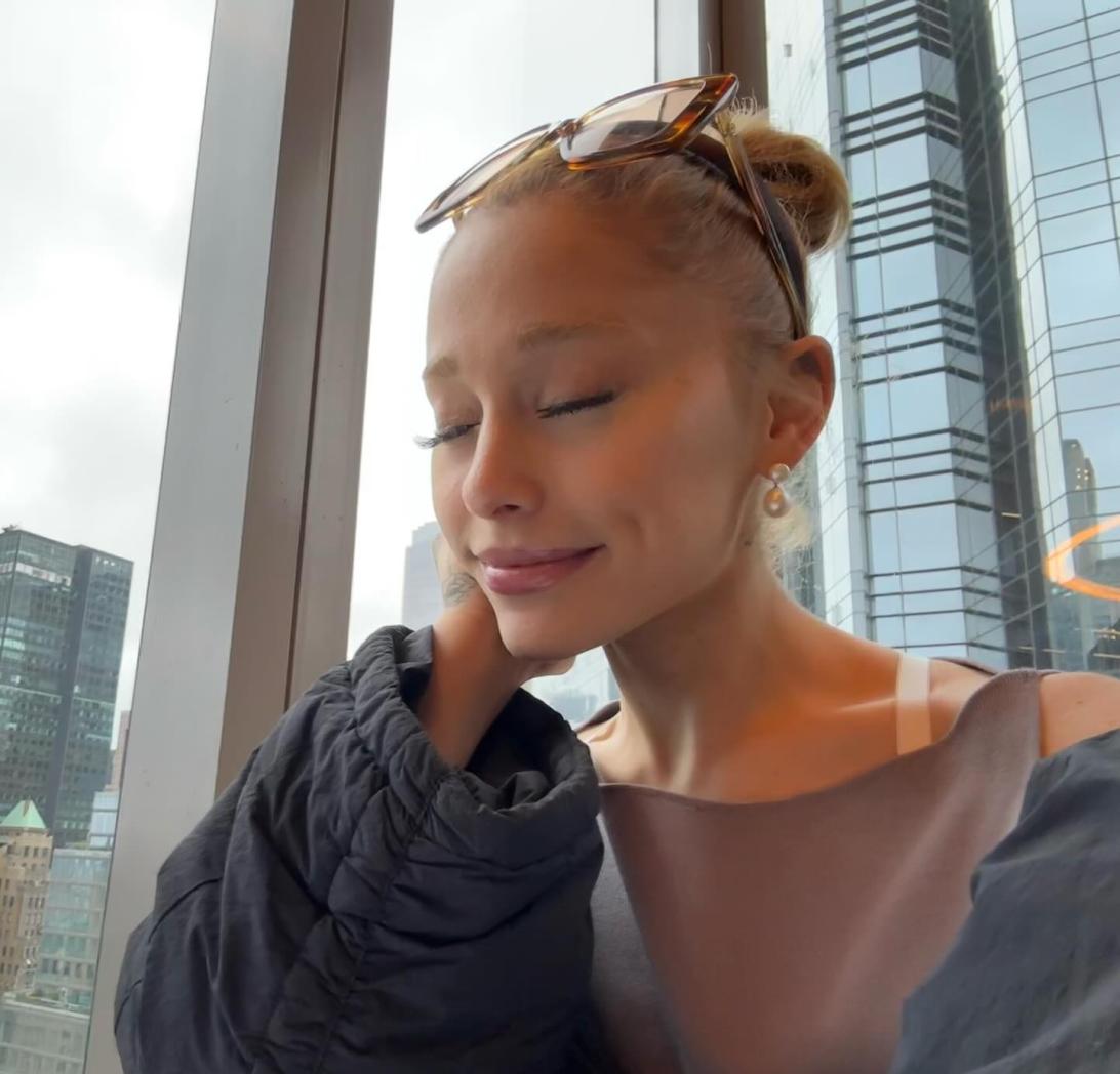 Ариана Гранде с закрытыми глазами, в темной куртке, сидит у окна с видом на городские здания.