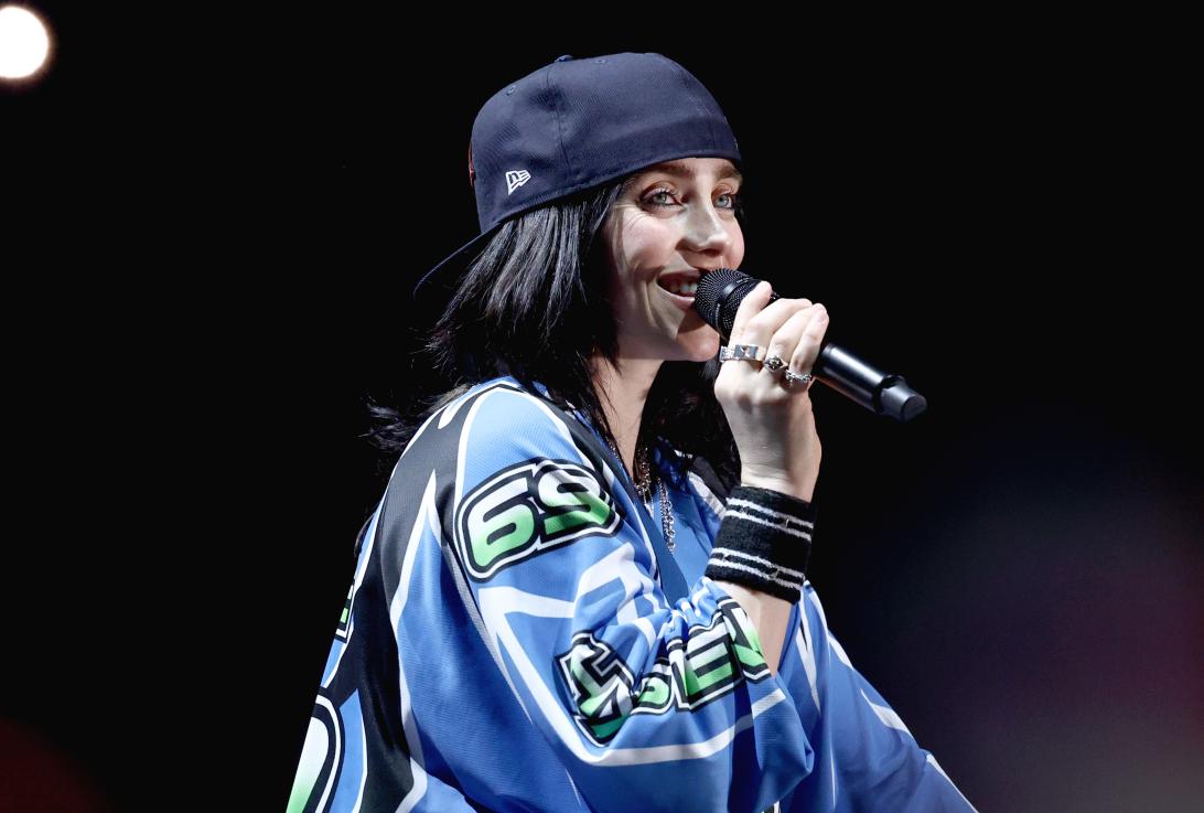 Билли Айлиш выступает на сцене в синей футболке и черной бейсболке, держа микрофон.