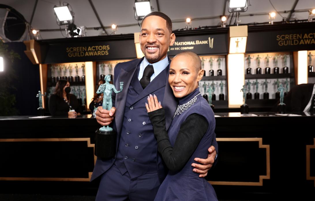 Уилл Смит с улыбкой держит награду, а Джада Пинкетт Смит, улыбаясь, нежно обнимает его.