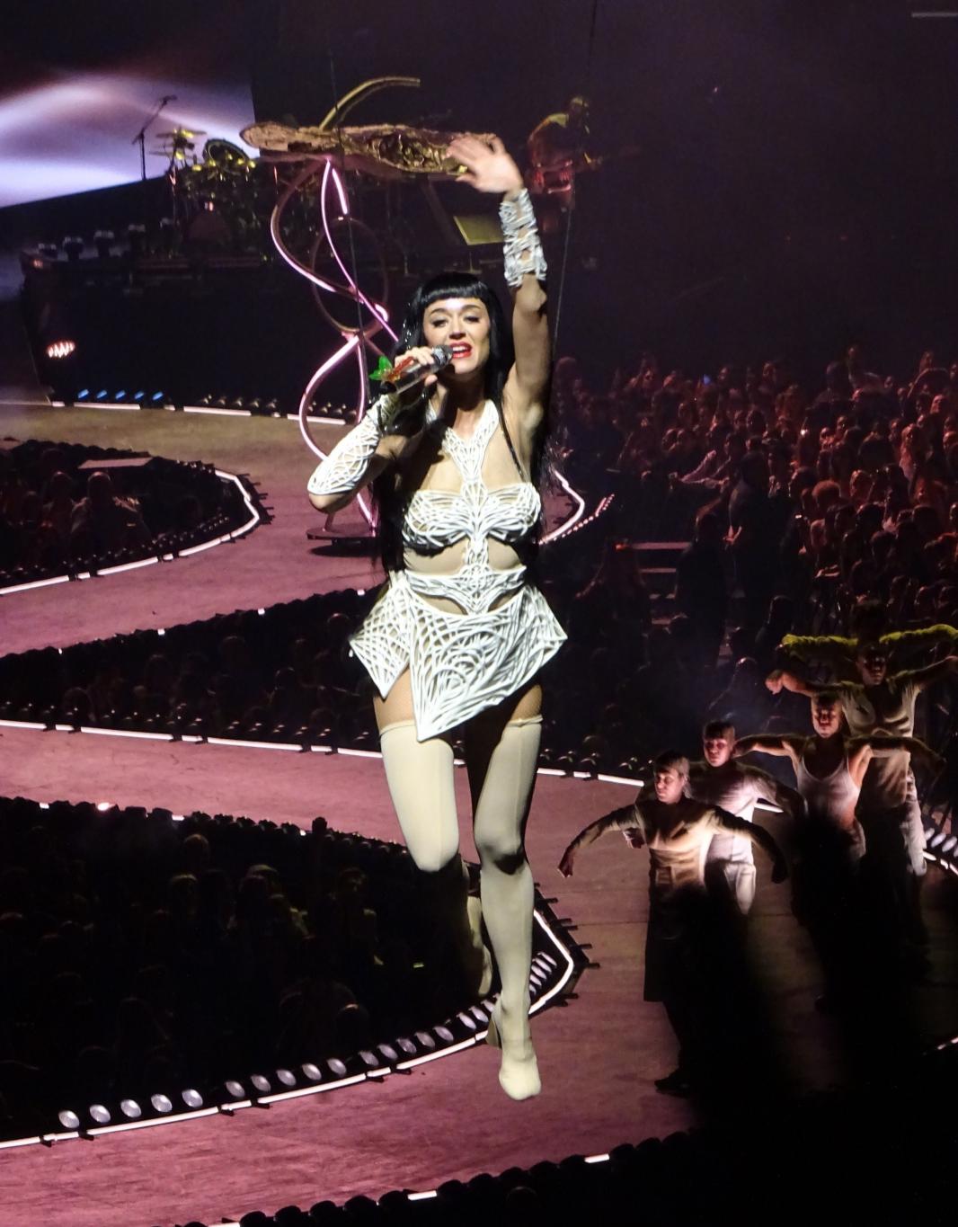 Кэти Перри выступает в Будапеште на своем туре "Lifetimes" в белом наряде, с микрофоном, окруженная толпой и танцорами.