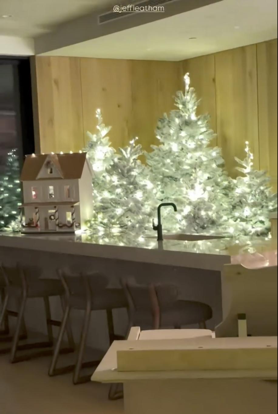 На кухонной столешнице — миниатюрный домик, а за ним — несколько заснеженных елочек, подсвеченных теплым белым светом.