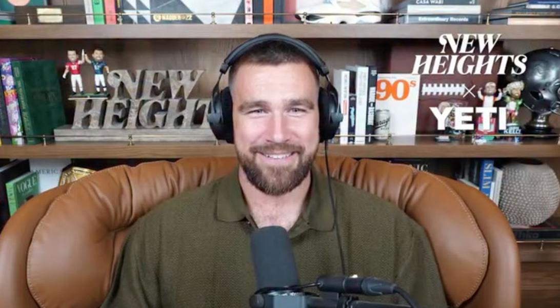 Трэвис Келси улыбается в наушниках и зеленом поло, сидя в кожаном кресле перед книжным шкафом с атрибутикой подкаста "New Heights".