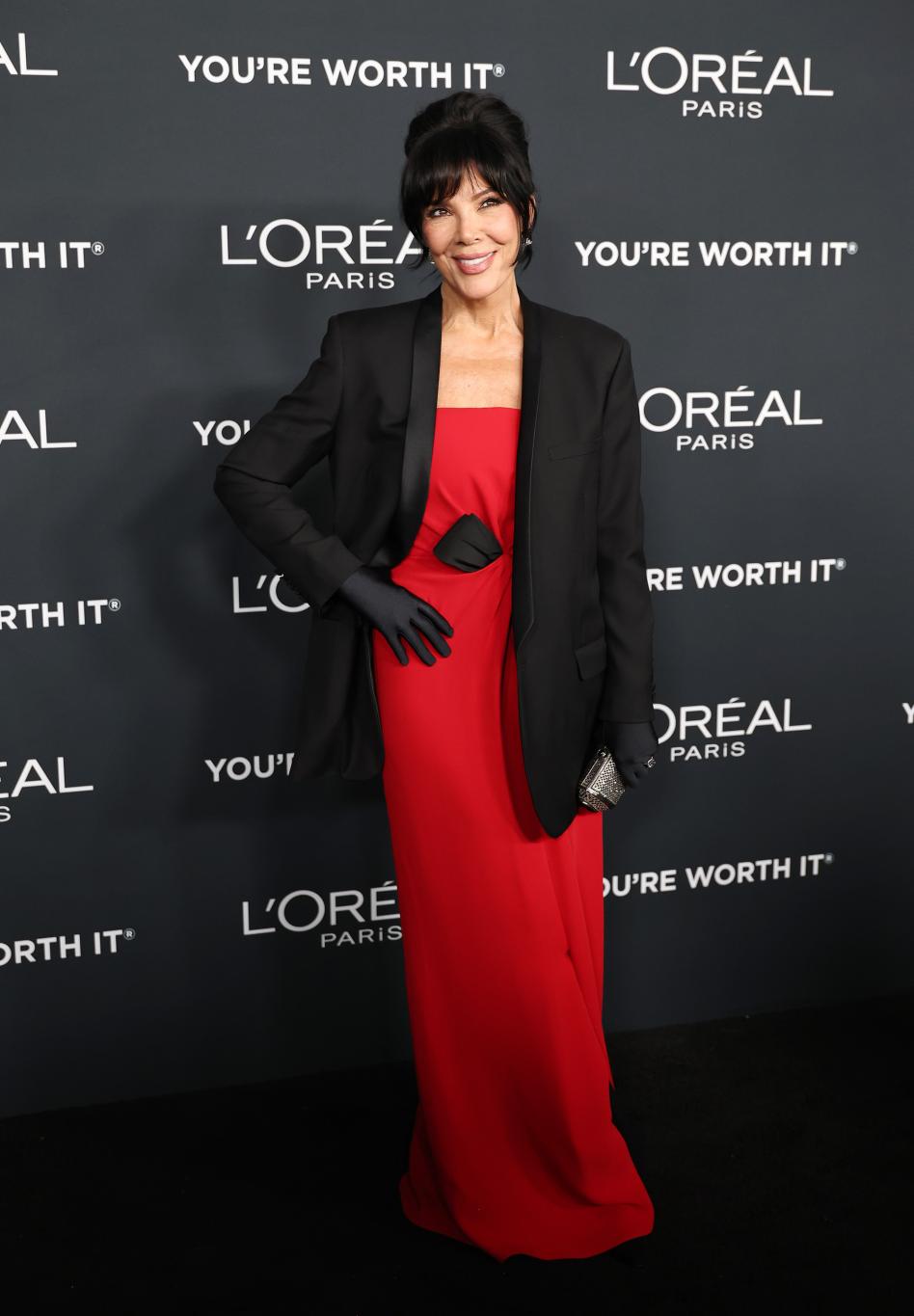 Крис Дженнер в алом платье, черном блейзере и черных перчатках позирует на фоне буквы L.