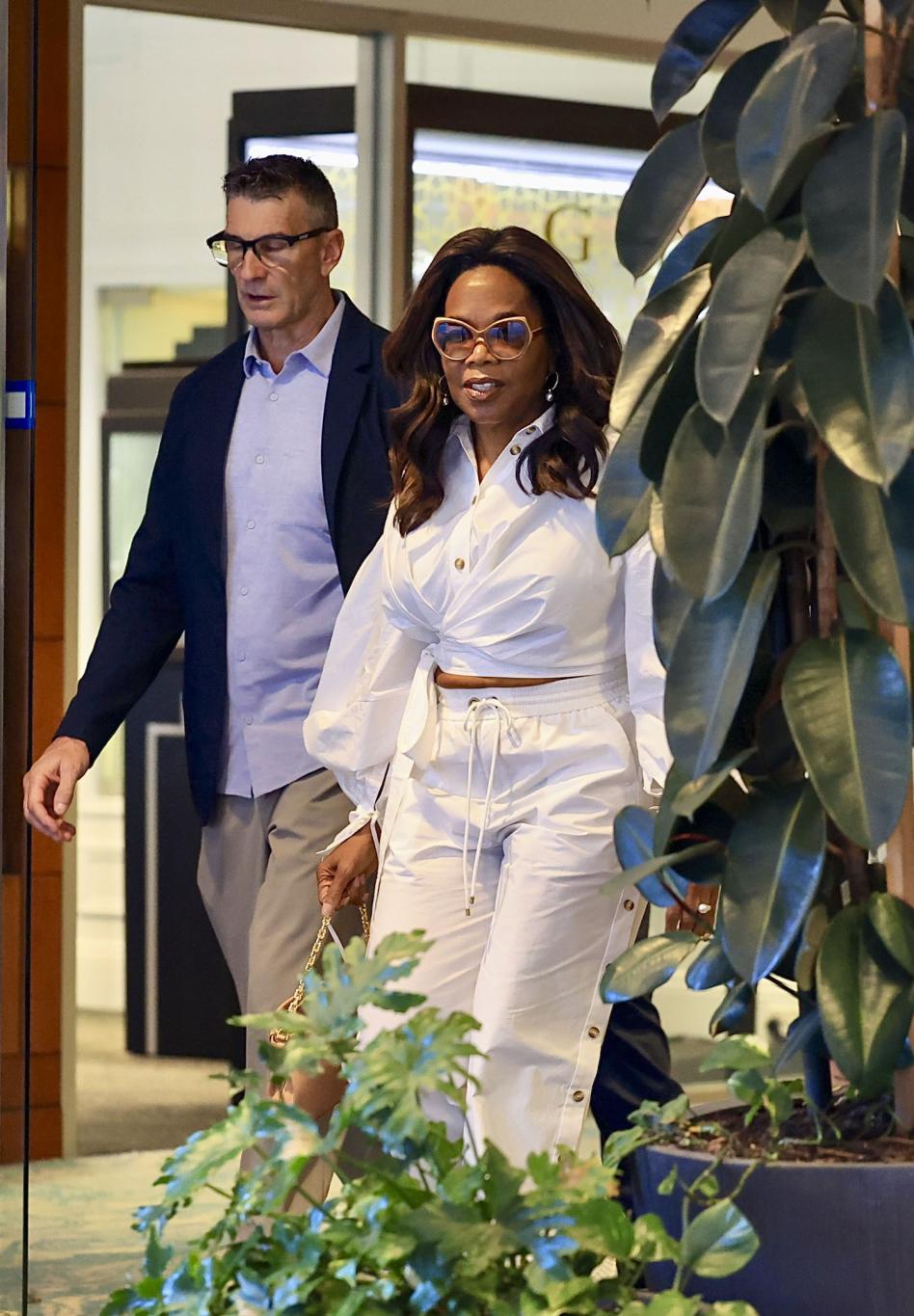 Опра Уинфри в белом кроп-топе, брюках с завышенной талией и тонированных очках, на фоне мужчины.