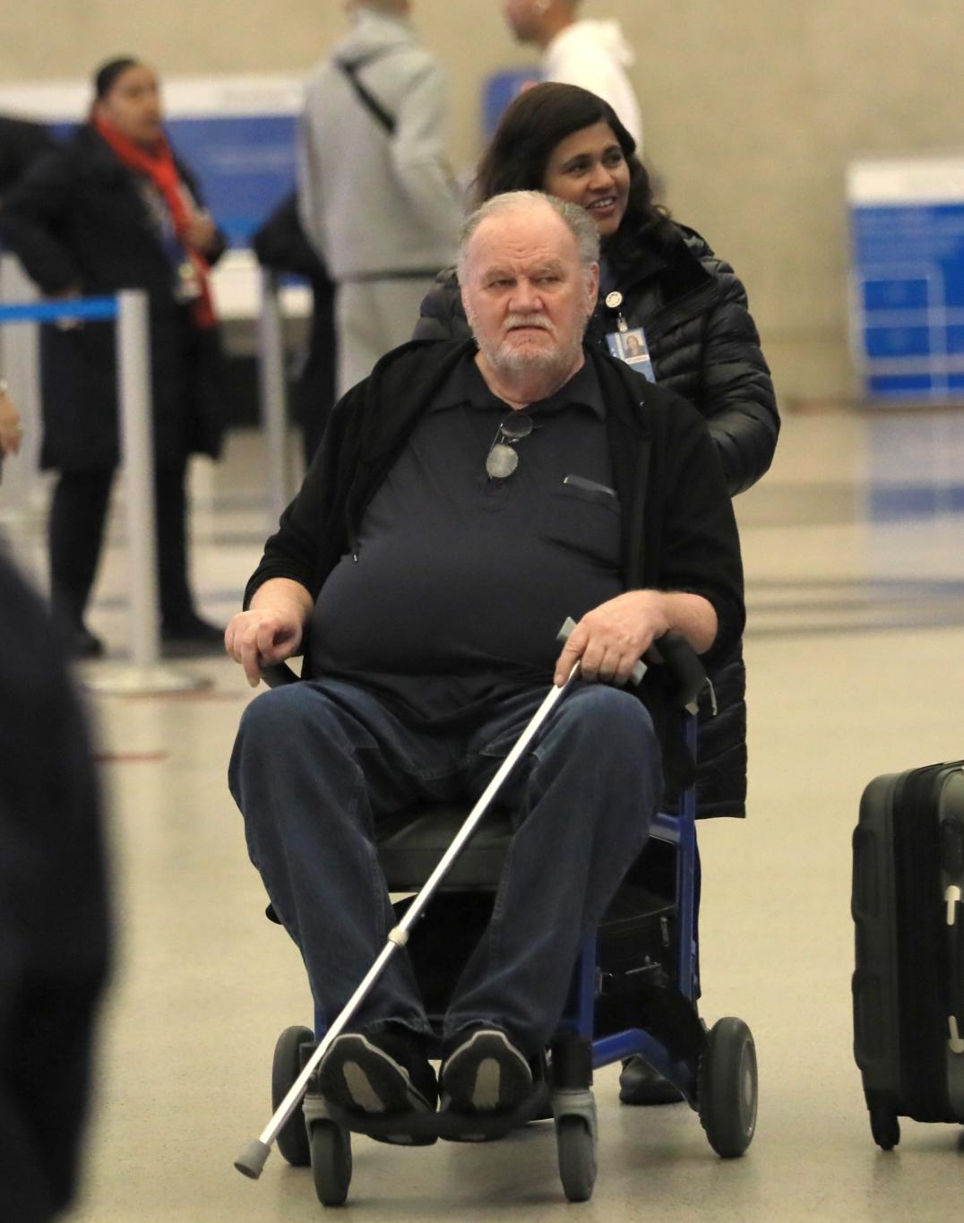Женщина везет Томаса Маркла в инвалидной коляске, с тростью, по аэропорту.