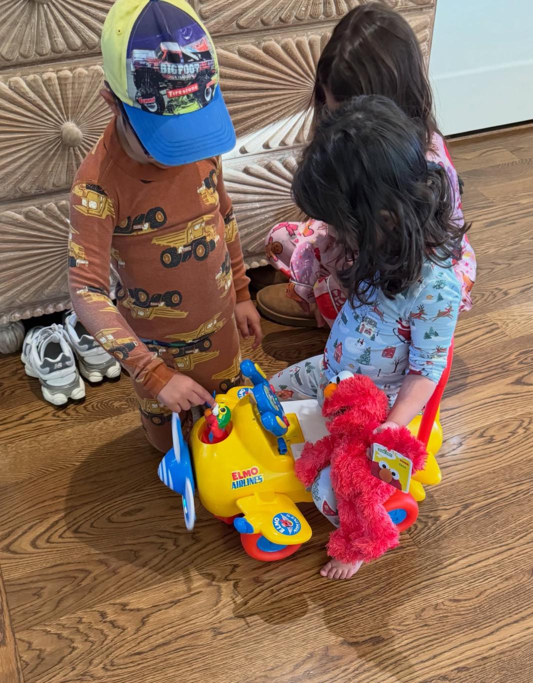 Трое детей играют на деревянном полу с желтым самолетиком Elmo Airlines и красной куклой Elmo.