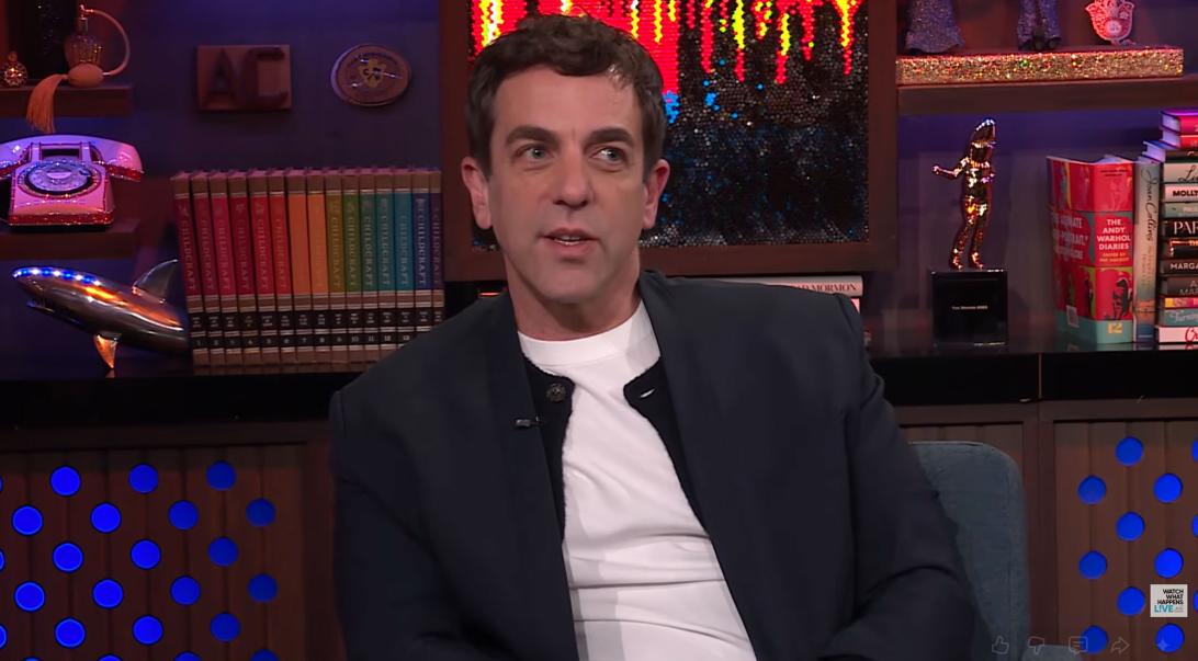 Эмджей Новак на синем диване в белой рубашке под черной курткой.