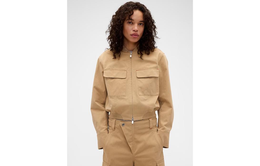 Женщина в бежевой куртке на молнии с четырьмя карманами и бежевых брюках с уникальной асимметричной пуговицей.