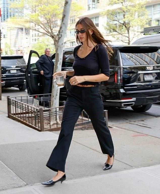 Основательница Rhode, 29 лет, пила кофе из фирменной кружки, прогуливаясь по тротуару в черном наряде.