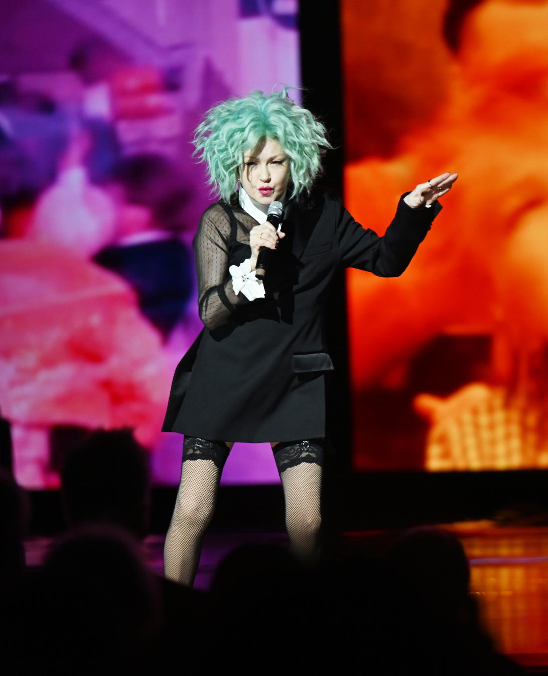 Синди Лопер в черном блейзере, сетчатом топе и чулках, с бирюзовыми кудрями, поет в микрофон.