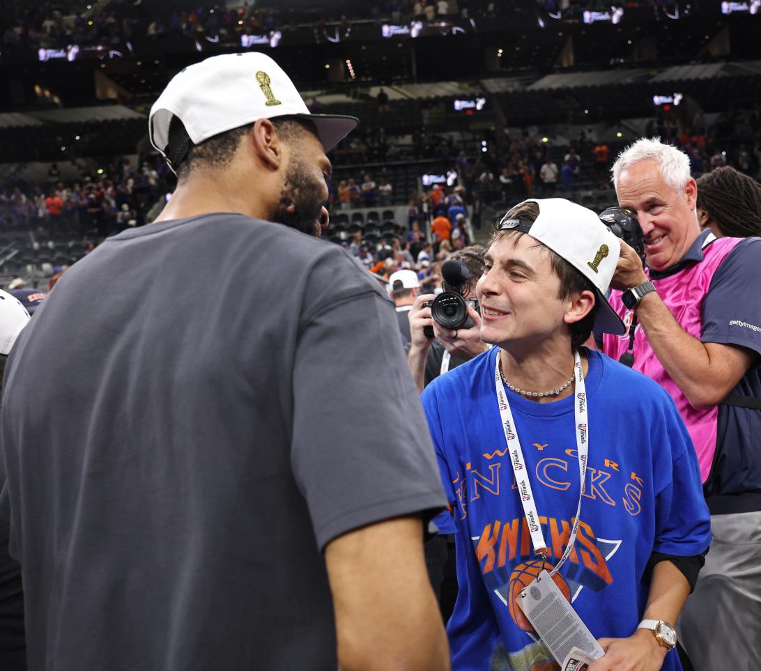 Тимоте Шаламе улыбается в футболке Knicks и чемпионской кепке, общаясь с игроком.